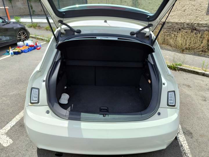 Coffre ouvert et vide d’une Audi A1 blanche sur un parking, matériel de nettoyage posé au sol à l’arrière-plan