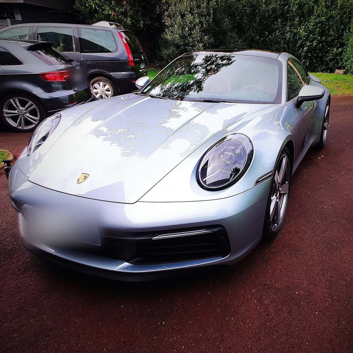 Porsche 911 gris argent vue de trois quarts avant, carrosserie brillante et phares ronds, garée dans une allée